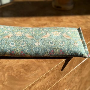 May include: A rectangular, patterned pillow with a teal background and a floral and bird design. The pillow is filled and has a soft, textured appearance. The design includes white flowers, birds, and red berries.