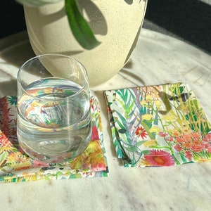 May include: Two square fabric coasters with a floral design. One coaster has a clear glass of water on top. The coasters feature a colorful watercolor pattern with flowers in shades of red, yellow, and green. A white vase is in the background.