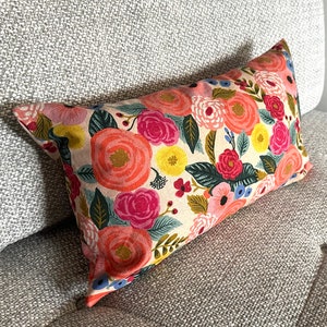 May include: Rectangular decorative pillow with a floral pattern. The pillow features a cream-colored background with large, colorful flowers in shades of pink, orange, yellow, and blue, with green leaves. The pillow is resting on a gray upholstered couch.