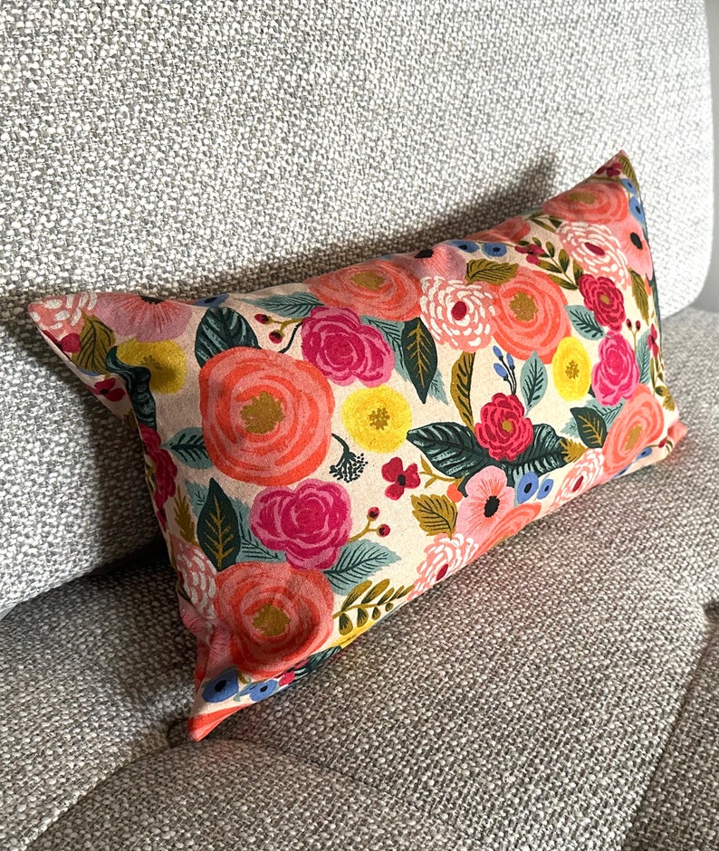 May include: Rectangular decorative pillow with a floral pattern. The pillow features a cream-colored background with large, colorful flowers in shades of pink, orange, yellow, and blue, with green leaves. The pillow is resting on a gray upholstered couch.