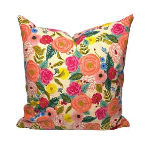 May include: A square decorative pillow with a cream-colored background and a vibrant floral pattern. The design features large, colorful flowers in shades of coral, pink, red, yellow, and blue, along with green leaves. The pillow is a soft, inviting home decor item.