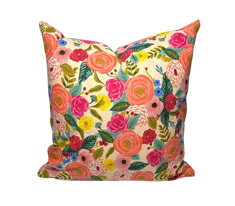 May include: A square decorative pillow with a cream-colored background and a vibrant floral pattern. The design features large, colorful flowers in shades of coral, pink, red, yellow, and blue, along with green leaves. The pillow is a soft, inviting home decor item.