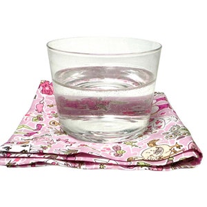 May include: A clear glass tumbler filled with water resting on a folded pink fabric napkin. The napkin features a repeating pattern of various small illustrations including animals, flowers, and objects in shades of pink, green, and white.
