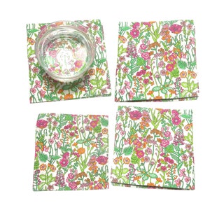 Puede incluir: Cuatro posavasos cuadrados con un estampado floral en rosa, naranja y verde sobre fondo blanco. Un posavasos con un vaso transparente encima. Los posavasos son adecuados para bebidas.