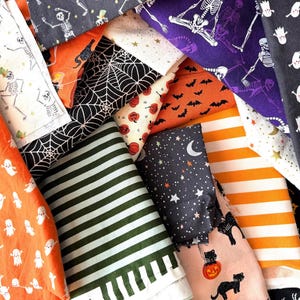 May include: A collection of folded cotton fabrics featuring Halloween-themed patterns including white ghosts on orange, black spiderwebs, skeletons on purple, orange pumpkins, black bats, green stripes, stars, moons, and black cats on pink.