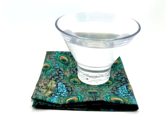 May include: A clear plastic cup sits on a folded square coaster. The coaster features a peacock feather design in shades of teal, green, and gold. The background is white.