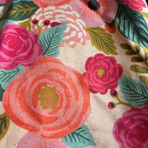 May include: Close-up of a floral fabric featuring large, stylized roses in shades of pink and coral. The design includes green leaves and smaller flowers in pink and blue, set against a cream-colored background.