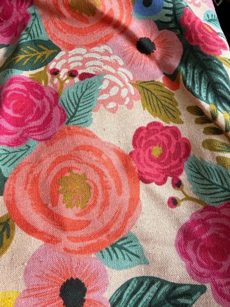 May include: Close-up of a floral fabric featuring large, stylized roses in shades of pink and coral. The design includes green leaves and smaller flowers in pink and blue, set against a cream-colored background.