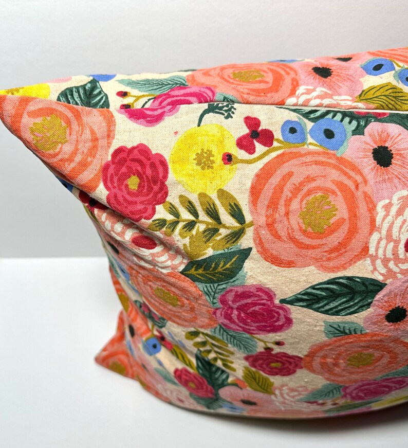 May include: A decorative pillow with a floral pattern. The pillow features large, colorful flowers in shades of pink, orange, yellow, and blue against a cream-colored background. Green leaves and stems are also visible.