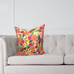 May include: A decorative throw pillow with a floral pattern featuring pink, orange, yellow, and blue flowers on a cream background. The pillow is placed on a white tufted sofa, adding a touch of color to the neutral setting.