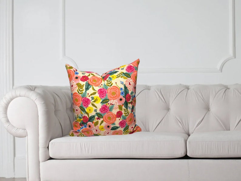 May include: A decorative throw pillow with a floral pattern featuring pink, orange, yellow, and blue flowers on a cream background. The pillow is placed on a white tufted sofa, adding a touch of color to the neutral setting.