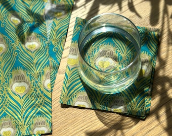 May include: A clear glass of water sits on a teal and gold peacock feather patterned coaster. The coaster and matching fabric are on a light wood surface. The design features teal, gold, and brown colors.