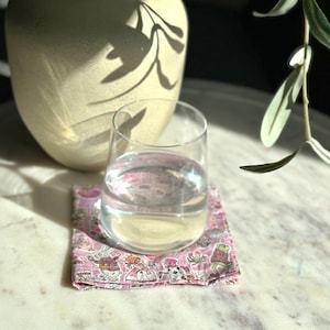 Könnte beinhalten: Ein Glas Wasser steht auf einem rosa-weiß gemusterten Untersetzer. Der Untersetzer hat ein sich wiederholendes Design von Cartoonfiguren. Im Hintergrund befindet sich eine beige Vase und rechts oben ein Zweig mit grünen Blättern.