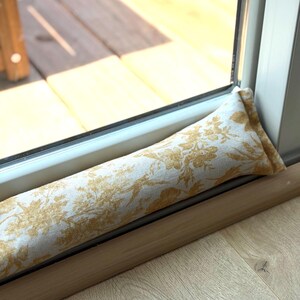 Op de afbeelding: Een lange, cilindrische tochtstopper met een goud- en wit bloemmotief. De stof is geplaatst tegen een raamkozijn en een houten vloer. De tochtstopper is ontworpen om tocht te blokkeren en de energie-efficiëntie te verbeteren.