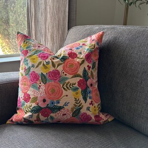 May include: A decorative throw pillow with a floral pattern. The pillow features large, colorful flowers in shades of pink, orange, and yellow, set against a beige background. Green leaves and accents complete the design. The pillow is placed on a gray sofa.