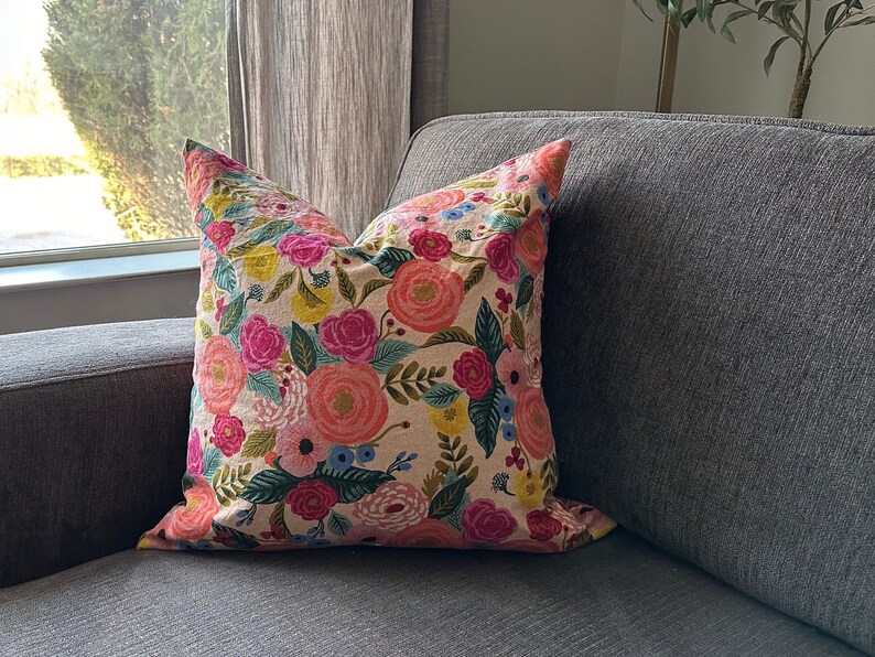 May include: A decorative throw pillow with a floral pattern. The pillow features large, colorful flowers in shades of pink, orange, and yellow, set against a beige background. Green leaves and accents complete the design. The pillow is placed on a gray sofa.