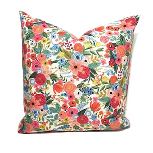 May include: A square decorative pillow with a vibrant floral pattern. The pillow features a white background covered in a colorful array of flowers, including red, pink, orange, yellow, and blue blooms, with green foliage. The pillow is a home decor item.