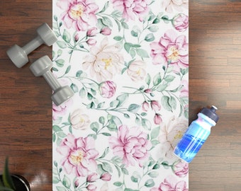 May include: A white yoga mat with a floral pattern featuring pink and white flowers and green leaves. The mat is on a wooden floor with a water bottle and two dumbbells.