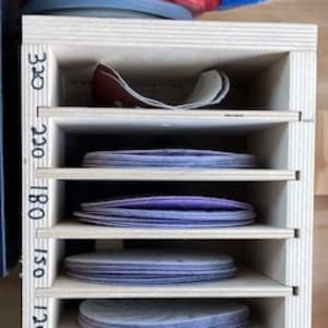 Puede incluir: Una lijadora eléctrica roja y negra con un mango azul está colocada encima de un organizador de madera. El organizador tiene varios estantes llenos de discos de lijado circulares de diferentes granos, etiquetados de 80 a 320.