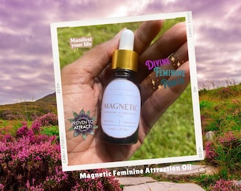 Puede incluir: Una botella de vidrio con una tapa dorada y una etiqueta que dice "Magnetic Feminine Attraction Oil". La botella se sostiene en una mano contra un fondo de cielo nublado y colinas verdes.