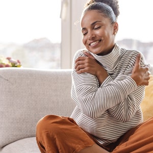 May include: A woman sitting on a couch with her arms wrapped around herself, smiling with her eyes closed. She is wearing a white and black striped sweater and brown pants.