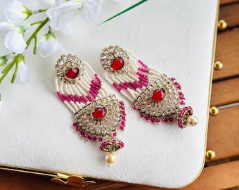 May include: A pair of ornate earrings with a red gemstone center and a pearl drop. The earrings feature a gold-colored filigree design with clear stones and white beaded strands. The design includes a zig-zag pattern in magenta beads.