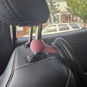 May include: A pink car headrest hook is attached to a black car seat. The hook is designed to hold items and is positioned near the headrest. A black, textured item is attached to the hook.