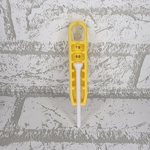 May include: A yellow golf tee holder with a white tee. The holder has the numbers 60 and 70 printed on it. The holder is made of plastic and has a loop at the top.