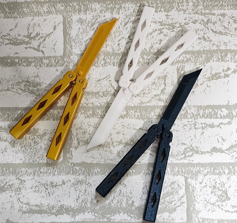 Puede incluir: Tres navajas mariposa, una dorada, una blanca y una negra. Las navajas est&aacute;n cerradas y dispuestas sobre un fondo de ladrillos blancos.