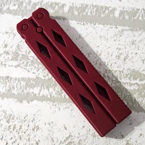 Puede incluir: Un cuchillo mariposa de metal rojo con un patr&oacute;n de diamante en el mango.