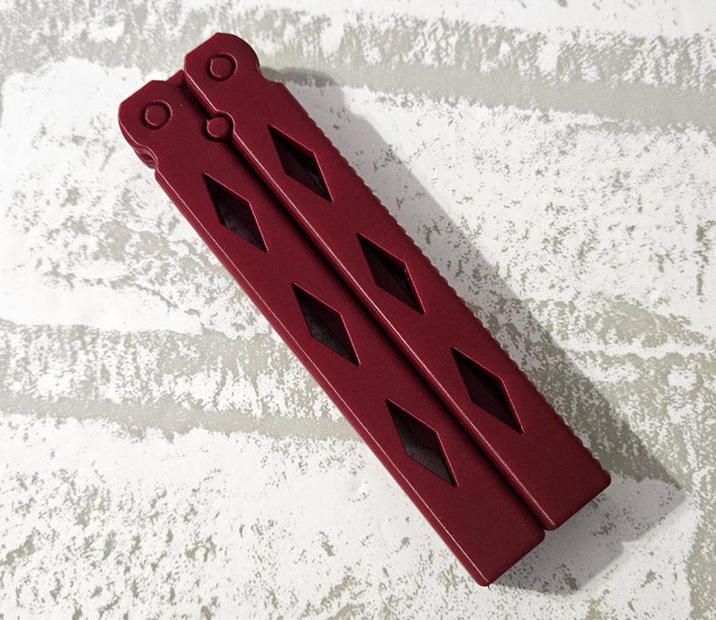 Puede incluir: Un cuchillo mariposa de metal rojo con un patr&oacute;n de diamante en el mango.