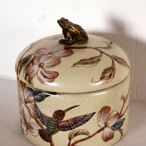 May include: A cream-coloured ceramic jar with a lid, decorated with a hummingbird and floral pattern. The lid is topped with a bronze frog figurine. The jar has a crackle glaze finish.
