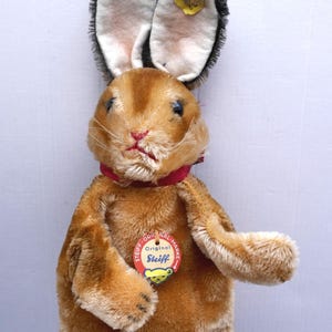 Könnte beinhalten: Ein Vintage-Steiff-Plüschkaninchen mit braunem Fell, langen Ohren mit schwarzen Spitzen und einem roten Schleifenkragen. Das Spielzeug hat ein Steiff-Etikett. Das Kaninchen ist ein klassisches Sammlerstück.