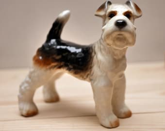 Puede incluir: Una figurita de cerámica blanca, negra y marrón de un fox terrier de pelo duro. El perro está de pie con la cabeza ligeramente inclinada hacia un lado.