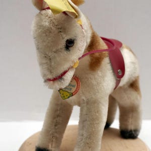 Op de afbeelding: Een wit en bruin knuffelpaard met een rood zadel en een geel label met de tekst "Original Steiff" met een teddybeerlogo. Het paard staat op een houten voet.
