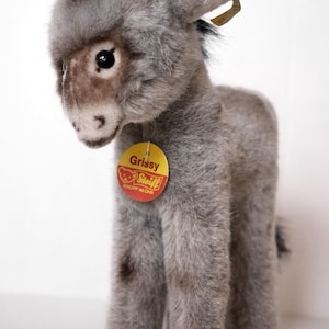 Puede incluir: Un juguete de peluche gris y marrón en forma de burro con pelo negro en la cabeza. El juguete tiene una etiqueta que dice "Grissy" y "Steiff Knopf im Ohr".
