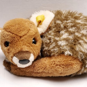 Puede incluir: Un peluche de cervatillo en posición encogida. El cervatillo tiene pelaje marrón con manchas blancas en la espalda y una cabeza marrón claro. Tiene ojos negros, un hocico blanco y orejas color crema. Se adjunta una cinta amarilla.