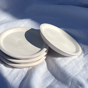 Könnte beinhalten: Ein Stapel von sechs cremeweißen, runden Keramikplatten. Eine Platte ist separat, leicht angewinkelt, um den Rand zu zeigen. Die Teller haben ein schlichtes, minimalistisches Design mit leicht erhöhtem Rand. Die Teller liegen auf einer weißen, strukturierten Oberfläche.
