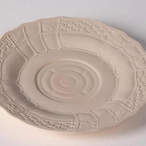 May include: A cream-coloured ceramic saucer with a scalloped edge and embossed geometric and floral patterns. The centre has concentric circles. The saucer is approximately 15 cm in diameter.