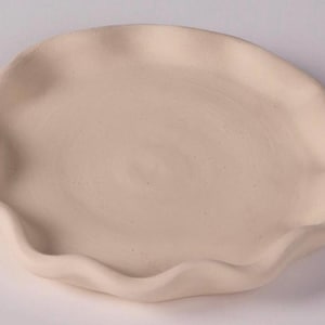 Puede incluir: Un plato de cerámica hecho a mano, de color blanquecino, con un borde ondulado y orgánico. El plato tiene un diseño poco profundo y circular y un acabado liso y mate. Este plato decorativo es adecuado para servir alimentos o como objeto decorativo.