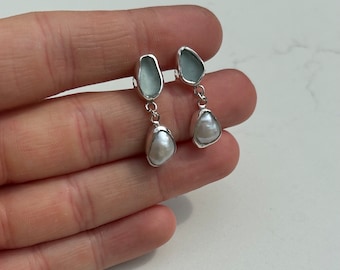 May include: Dangle earrings featuring a light blue, irregular-shaped stone at the top and a white, teardrop-shaped pearl at the bottom. The earrings are connected by a silver-colored metal. The earrings are displayed against a white background.