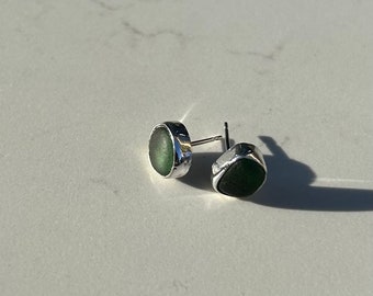 Puede incluir: Pendientes de botón con piedras verdes oscuras de forma irregular, engastadas en metal plateado. Los pendientes se muestran sobre una superficie blanca. El diseño es sencillo y elegante.