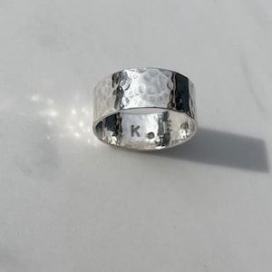 May include: A wide, hammered silver ring. The ring has a textured surface with small indentations. The inside of the ring has the letters "K.O.E." stamped into the metal. A simple, elegant piece of jewellery. The ring is a simple, yet elegant piece of jewellery.