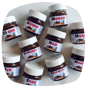 Peut inclure: Dix petits pots en verre avec des couvercles blancs remplis de pâte à tartiner au chocolat. Chaque pot a une étiquette différente avec un nom écrit en noir et blanc. Les noms incluent "Alyssa", "Maeva", "Noa", "Mate", "SoHappy", "Maya", "Enzo", "Bebe arrive", et "Maeli".