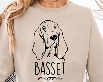 Może przedstawiać: Beżowa bluza z czarną grafiką przedstawiającą głowę Basset Hound i napisem "BASSET mom". Bluza ma długie rękawy i okrągły dekolt.