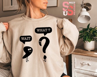 Op de afbeelding: Een beige sweatshirt met een zwarte afbeelding van een komma en een vraagteken met gezichten. De komma zegt "WAIT" en het vraagteken zegt "WHAT?"