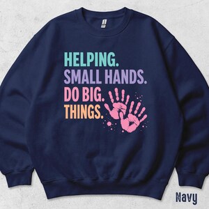 May include: A navy blue crewneck sweatshirt featuring the text HELPING. SMALL HANDS. DO BIG. THINGS. in teal, purple, pink, and orange lettering. To the right of the text are two pink handprints with small paint splatters around them.
