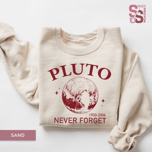 May include: A beige sweatshirt with a red graphic print of Pluto, a planet with stars, and the text "Pluto 1930-2006 Never Forget".