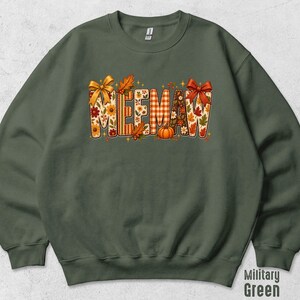 May include: A military green crewneck sweatshirt featuring the text MEE-MAW in large block letters. The letters are filled with a fall-themed pattern of orange pumpkins, autumn leaves, sunflowers, and plaid, accented by two orange bows.