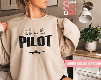May include: A beige sweatshirt with the text "dibs on the PILOT" in black lettering and a silhouette of an airplane. The sweatshirt is being worn by a person.